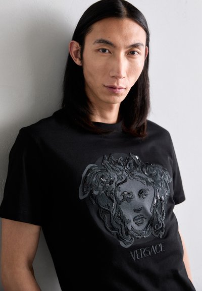Black cotton t-shirt featuring a textured black sequined design of a face, with the word "VERSACE" embroidered below in a matching color.