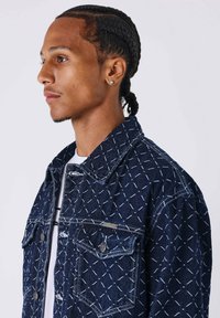 Chaqueta de mezclilla en azul oscuro con un patrón de costura blanca, con un collar clásico, dos bolsillos frontales y acentos de hardware en tono plateado.