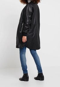 Zwarte leren lange jas met een relaxte pasvorm, voorzien van een zijrits zak, gedragen over blauwe jeans en zwarte enkellaarzen. Glad textuur.
