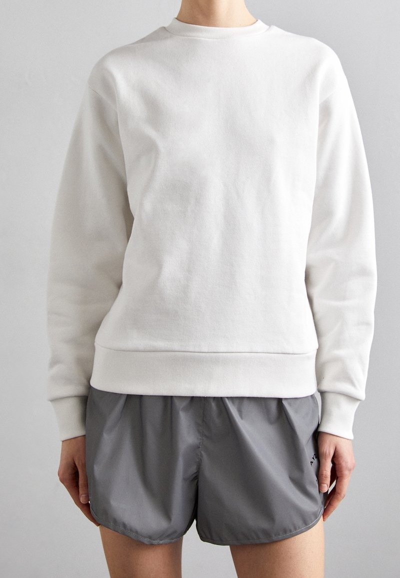 Personne portant un sweat-shirt blanc uni à manches longues et un short de sport gris ample, debout devant un fond neutre.