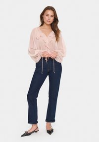 Blusa di chiffon rosa pallido con collo arricciato e polsini elastici, abbinata a jeans dritti blu scuro e ballerine nere a punta.