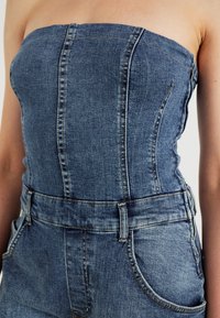 Denim overalls i medelblått. Utan axelremmar med strukturerad bodice, bälte i midjan och kurviga sidofickor. Distinkta sömnadsdetaljer.