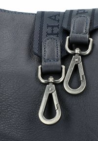 Ganchos de metal giratorios sujetos a correas de cuero negro y lazos de tela sobre la superficie rugosa de un bolso oscuro.