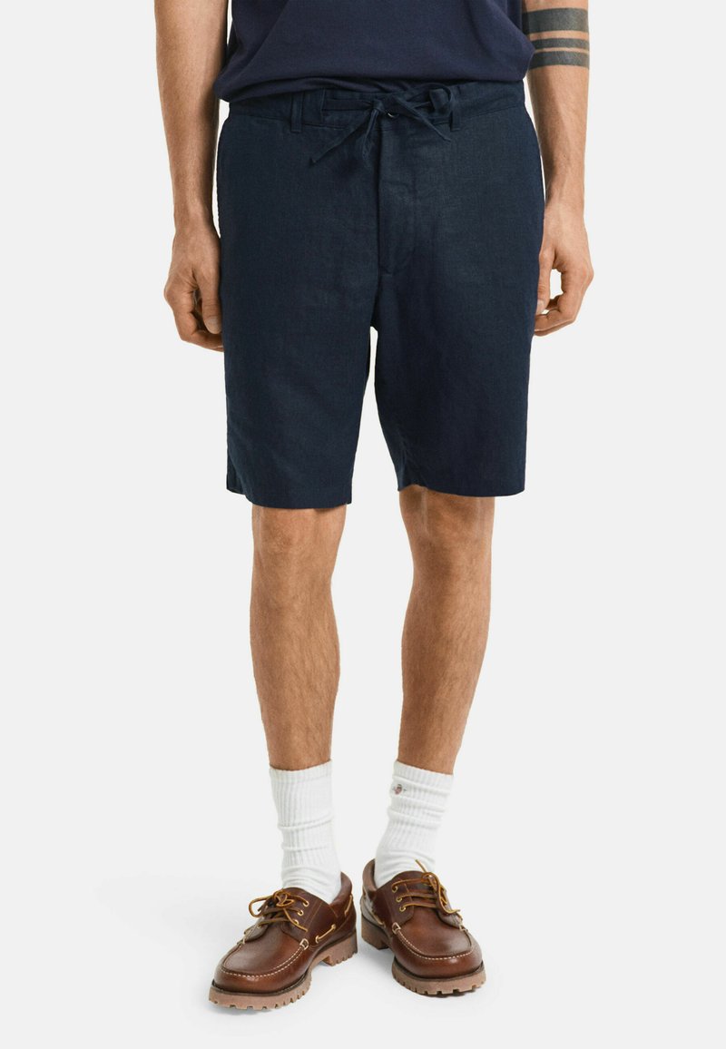 Marineblauwe linnen shorts met een verstelbare trekkoord taille, zijzakken en een relaxte pasvorm, gecombineerd met bruine schoenen en witte sokken.
