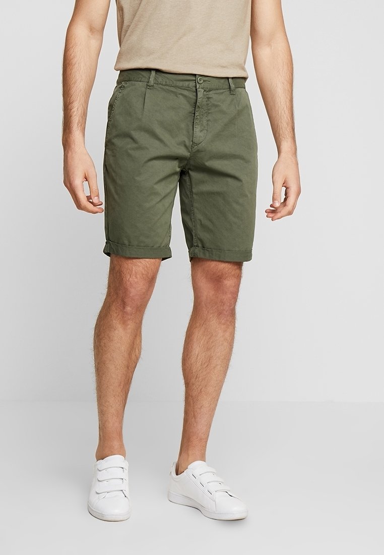 Homme portant un short vert olive retroussé, un t-shirt beige et des baskets blanches à scratch, debout devant un fond clair uni.