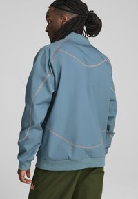 Gaiši zila jaka ar kontrastējošu bēškrāsas šuvēm, ar paceltu apkakli un ribotiem piedurknēm, kombinēta ar zaļām biksēm.