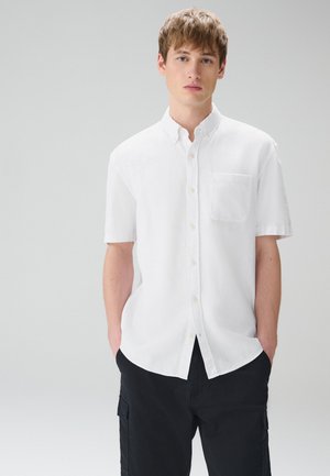 Noore mehenahe helepruuni juustega, kandes lühikeste varrukatega valget nööpidega särki ja musta pükse, seisab käed taskutes halli tausta ees.