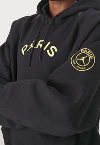 Sweat à capuche noir en tissu doux, avec un logo jaune "PARIS" sur la poitrine et un logo PSG circulaire sur la manche. Tissu texturé, capuche ajustable.
