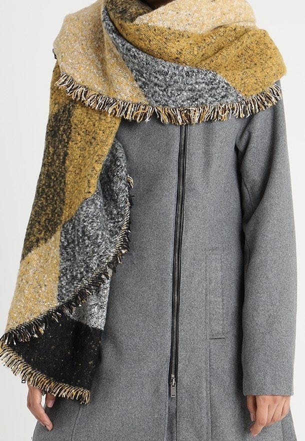 Écharpe épaisse en tricot moutarde, grise et noire avec des bords à franges portée par-dessus un manteau en laine gris à fermeture éclair.