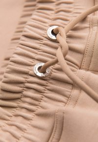 Ceinture élastique beige avec fronces cousues et un cordon assorti passant à travers des œillets métalliques sur le tissu.
