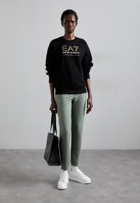 Zwarte sweatshirt met gouden branding, groene slim-fit broek, witte sneakers en een zwarte tote bag. Glad materiaal en een relaxte silhouet.