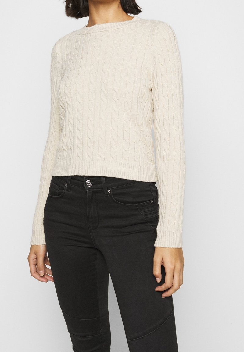 Maglione a treccia color crema con scollo rotondo e maniche lunghe, caratterizzato da orlo e polsini a coste. Abbinato a jeans neri aderenti.