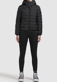 Czarna kurtka puchowa z zapięciem na zamek, teksturowanym wzorem fal i kapturem. Noszona z czarnymi legginsami i czarno-białymi butami sportowymi.
