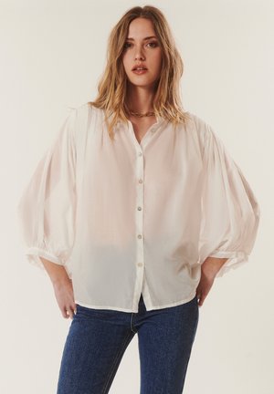 Femme avec un chemisier blanc ample boutonné avec des manches bouffantes et un jean bleu, debout les mains dans les poches devant un fond uni.