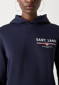 Námořnická modrá mikina s kapucí vyrobená z jemné tkaniny, s předním logem a textem "GANT 1949" a ikonou štítu, doplněná červenými a bílými pruhy.