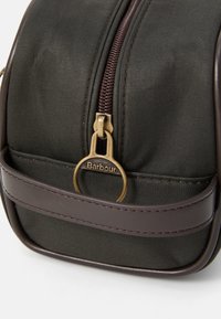 Close-up de uma bolsa Barbour verde escura mostrando o puxador de zipper de latão e o detalhe da alça de couro castanho em um fundo branco.