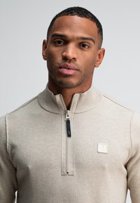 Beige ritssluitingtrui met een geribbelde kraag, voorzien van een klein vierkant logo op de borst en een contrasterende ritstrekkertoevoeging.