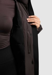 Schwarze Pufferjacke mit glattem Innenfutter, ausgestattet mit einem Druckknopfverschluss vorne und einer Seitentasche, teilweise offen gezeigt.