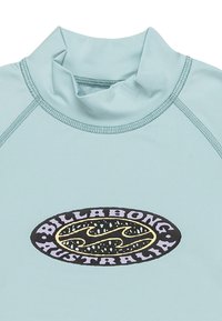 Ljusblå rash guard med hög krage, med en svart oval logotyp med gula detaljer som visar "BILLA BONG AUSTRALIA" i fet stil.