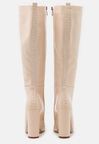 Botas de imitación de cuero beige hasta la rodilla con un acabado brillante, cordones detallados en la parte posterior y tacones de bloque grueso. Cierre con cremallera en el lado.