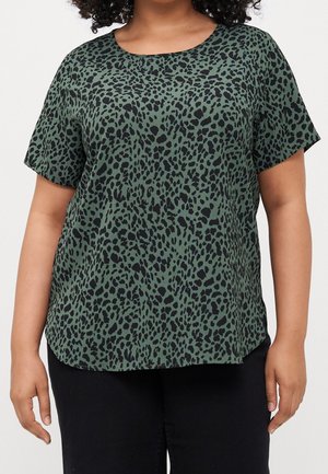 Kvinde iført en kortærmet grøn top med sort leopardprint og sorte bukser, stående mod en ensfarvet baggrund.