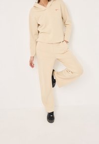 Femme portant un sweat à capuche beige et un pantalon large assorti, debout sur une jambe avec des baskets noires sur fond blanc.