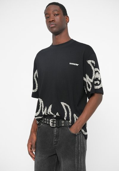 Must mustaga valgete kaunistustega, lühikeste varrukatega, ümmarguse kaelusega, koos hallide teksade ja studitud musta vööga.