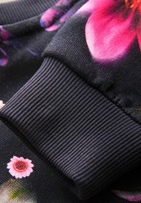 Mustast kangast dressipluusi varrukas, millel on sooniline tekstuur ja värvikad lillemustrid roosade ja lillade toonidega tumedal taustal.