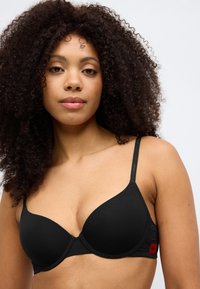 Fekete testhezálló melltartó sima anyagból, alátámasztó merevítővel és állítható pántokkal. Oldalán egy apró piros logó található részletként.