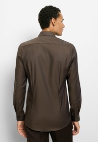 Braunes langärmliges Hemd mit strukturiertem Muster, Knopfmanschetten und spitzem Kragen, von hinten vor weißem Hintergrund gesehen.