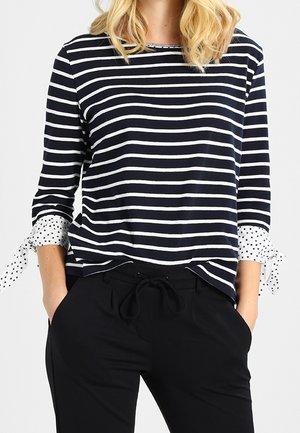 Femme portant un haut à manches longues rayé bleu marine et blanc avec des liens à pois aux poignets et un pantalon noir à cordon, les mains dans les poches.