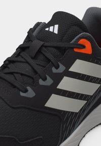 Μαύρο αθλητικό παπούτσι με λευκές ρίγες, μαύρα κορδόνια, υφή στη γλώσσα και πορτοκαλί λεπτομέρεια κοντά στην τρύπα των κορδονιών.