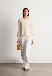 Crème cardigan met een gestructureerde weving, voorzien van ronde knopen. Gepaard met witte wijde broek en een kleine gele handtas. Glad schoeisel.