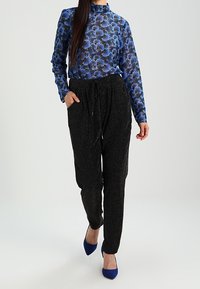 Långärmad blommig blus i blått med hög krage, matchad med svarta texturerade byxor. Modellen bär blå klackar. Enkelt och modernt plagg.