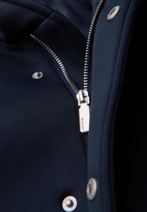 Marineblauwe jas met een zilveren rits, drukknoopsluitingen en een gestructureerde stof, met een zichtbaar logo aan de ritsloper.