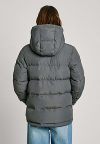 Giacca trapuntata grigia con cappuccio, caratterizzata da un design trapuntato e motivi di cuciture visibili. Il tessuto appare liscio e leggermente riflettente.