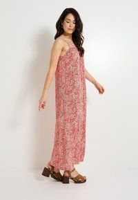 Robe maxi à pois rouges avec des fines bretelles et une coupe fluide, associée à des sandales à talons bruns avec des accents cloutés.