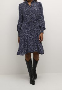 Abito a stampa floreale in blu navy scuro con fiori viola, maniche lunghe a sbuffo, colletto, vita con fiocco, gonna svasata, abbinato a stivali neri al ginocchio.