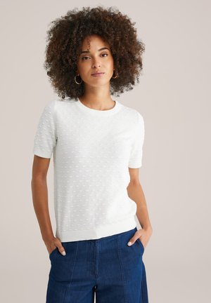 Young woman with curly hair wearing a textured white short-sleeve sweater and dark blue pants, hands in pockets, neutral background.