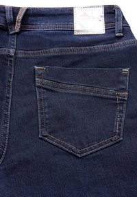 Jean en denim bleu foncé avec une poche arrière ornée de surpiqûres beiges et d'une étiquette rectangulaire à la taille. Texture lisse.