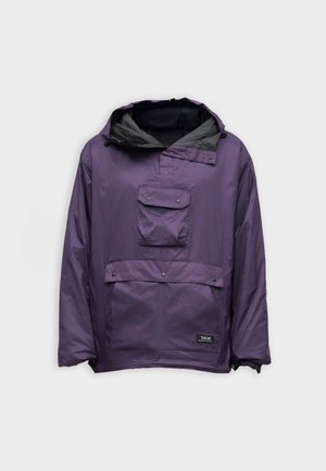 Jonge man die een losse paarse capuchontrui met zakken aan de voorkant en een zwarte broek draagt, staand tegen een eenvoudige lichte achtergrond.