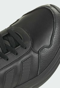 Gros plan d'une basket en cuir noire avec des bandes latérales texturées et des lacets noirs sur un fond clair.