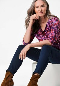 Lila und rot gemusterte Bluse mit einem drapierten Ausschnitt, kombiniert mit dunkelblauen Jeans und braunen Wildlederschnürstiefeln. Glatte Stofftextur.