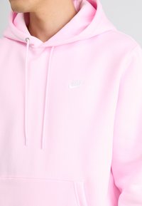 Lyse rosa Nike hettegenser med kengurulomme, snorhette og hvit logo på brystet. Mykt materiale med glatt tekstur.