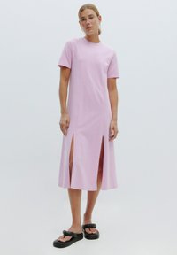 Kurzes pinkes Kleid mit kurzen Ärmeln, seitlichen Schlitzen und relaxed Fit, aus weichem Baumwollstoff. Getragen mit schwarzen Slide-Sandalen.