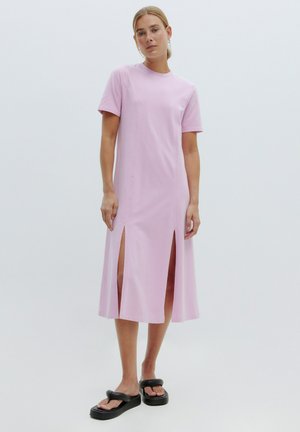 Robe courte rose avec des manches courtes, fentes sur les côtés et coupe décontractée, fabriquée en tissu de coton doux. Portée avec des sandales à glissière noires.