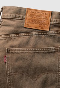 Brown denim jeans feature a leather patch with embossed branding and stitching details, including a red tag at the pocket.