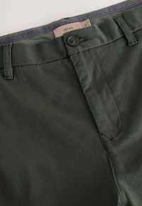 Pantalon vert foncé avec fermeture à boutons et passants pour ceinture, l'étiquette de la taille étant partiellement visible à l'intérieur.