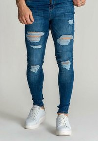Vaaleansiniset skinny-farkut, joissa on useita kulutettuja repeämiä. Pehmeä denimkangas, joka istuu tiukasti, yhdistettynä valkoisiin lenkkareihin.