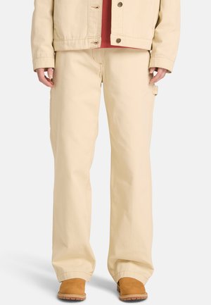 Pantaloni cargo beige, ampi, con tasche laterali, cuciture rinforzate e tessuto di cotone morbido. Abbinati a calzature marroni.